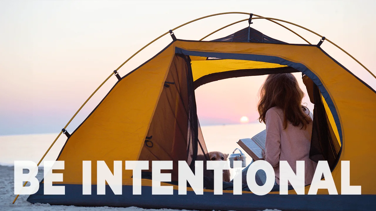 A serene scene of a woman reading inside a yellow tent by the beach during sunset with the bold words "BE INTENTIONAL" overlaying the bottom of the image.